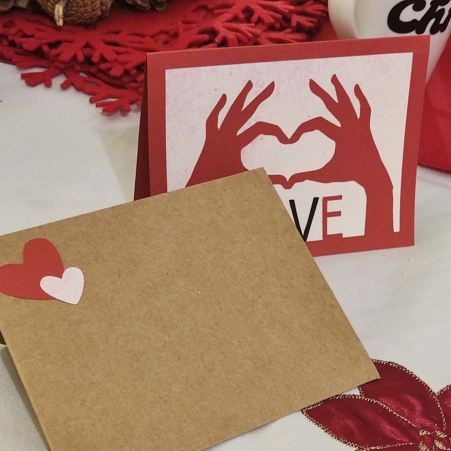 Handmade love card with heart hands design paired with a kraft envelope decorated with red and pink paper hearts.