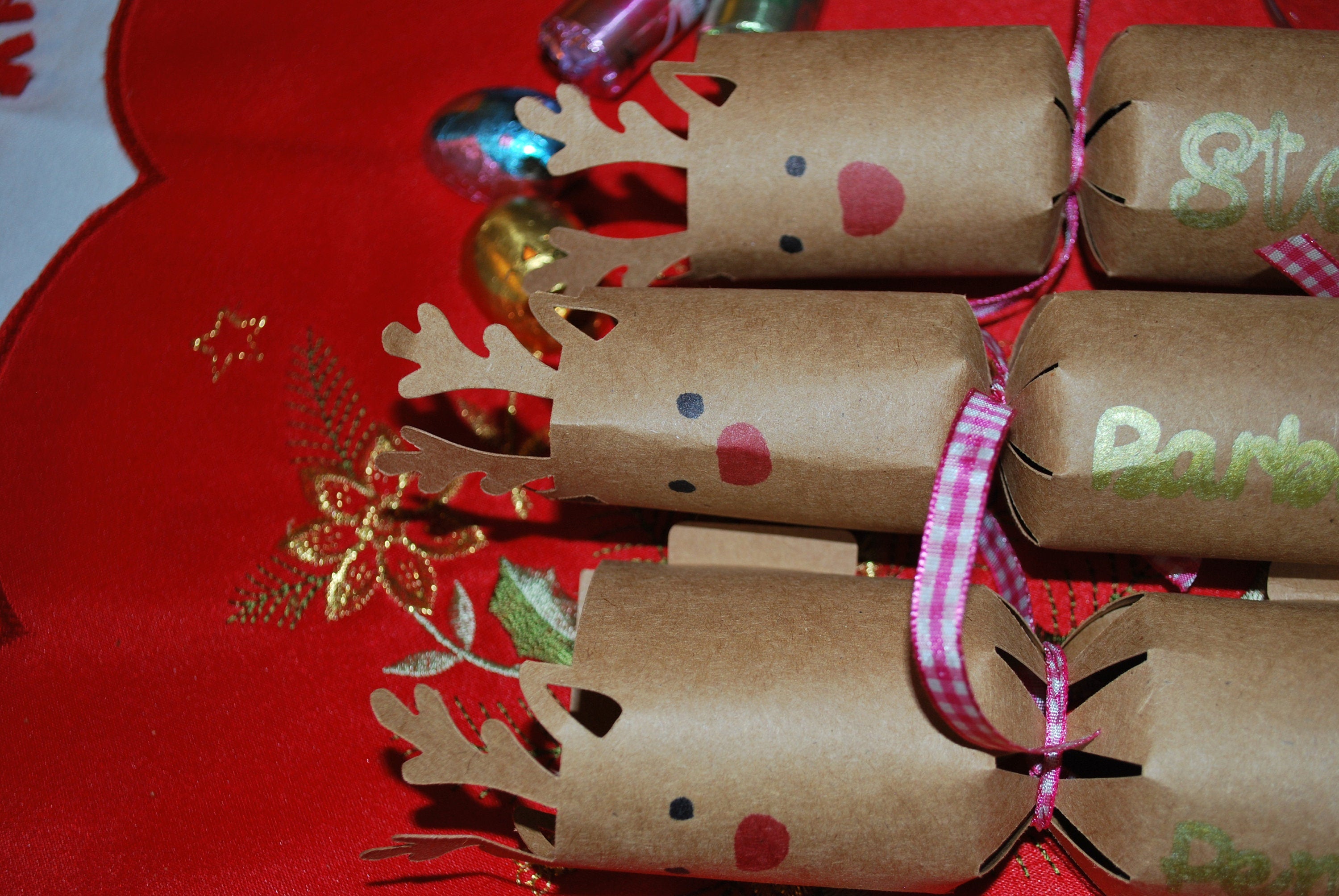 Group of handmade reindeer crackers with antlers, red noses, and pink gingham ribbons, personalised with names in gold foil, styled on festive red table linen.