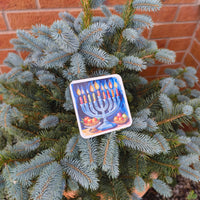Coaster with glowing menorah and striped candles, resting on evergreen tree branches outdoors