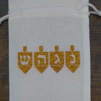 Handmade white drawstring hessian gift bag displayed on a dark wooden background. The front of the bag features four gold glitter dreidels, each with a Hebrew letter—Shin, Hey, Gimel, and Nun—representing the Hanukkah phrase “A great miracle happened there.” The festive design is cantered on the bag.