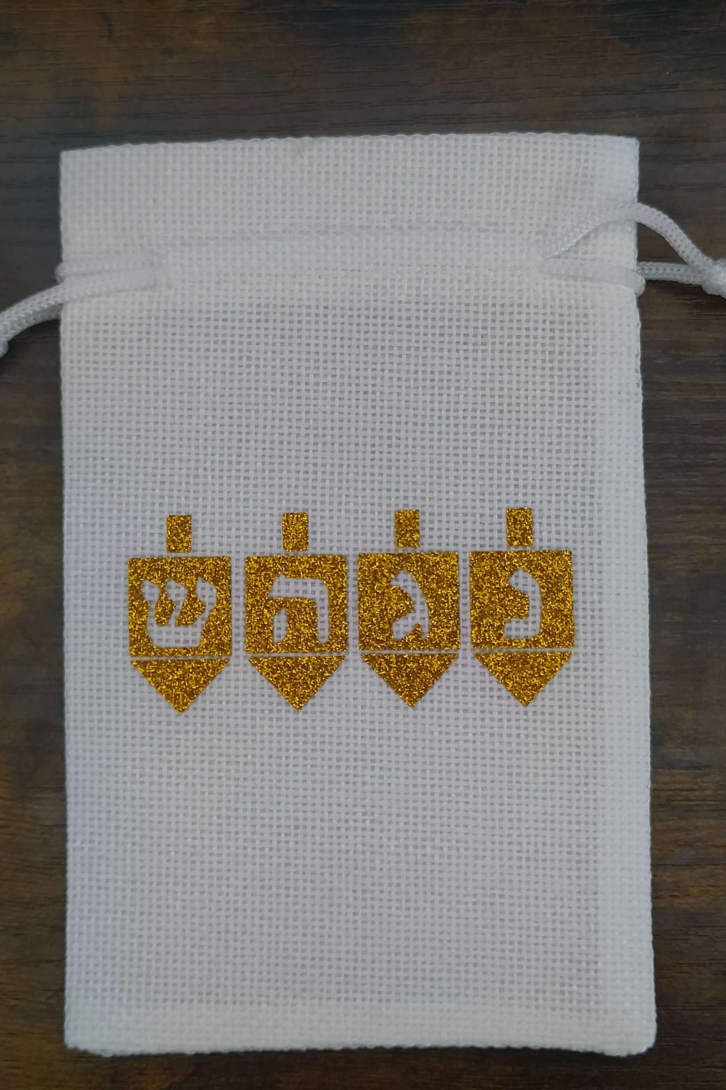 Handmade white drawstring hessian gift bag displayed on a dark wooden background. The front of the bag features four gold glitter dreidels, each with a Hebrew letter—Shin, Hey, Gimel, and Nun—representing the Hanukkah phrase “A great miracle happened there.” The festive design is cantered on the bag.