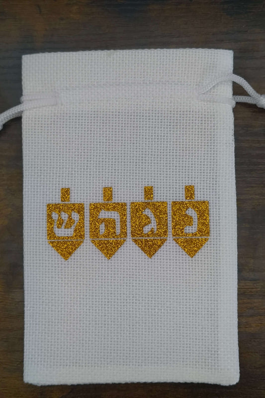 Handmade white drawstring hessian gift bag displayed on a dark wooden background. The front of the bag features four gold glitter dreidels, each with a Hebrew letter—Shin, Hey, Gimel, and Nun—representing the Hanukkah phrase “A great miracle happened there.” The festive design is cantered on the bag.