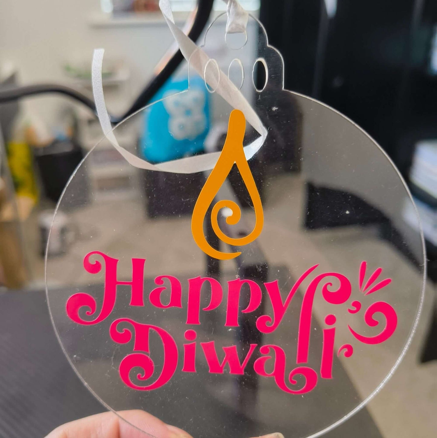 Clear acrylic Diwali ornament with pink “Happy Diwali” text and orange diya flame design, hung with white ribbon for festive home decor.