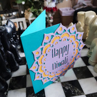 Handmade Diwali card with layered mandala design in purple, gold, and teal. Textured cardstock stands upright, showing bold festive colours.