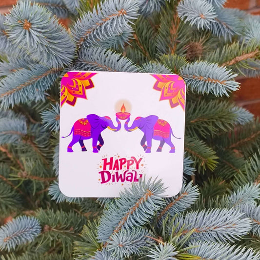 Close-up of a Happy Diwali coaster with two purple elephants and a glowing diya, resting on blue spruce branches.