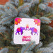 Happy Diwali coaster with two purple elephants and a central diya, surrounded by mandala motifs, photographed among blue spruce branches.