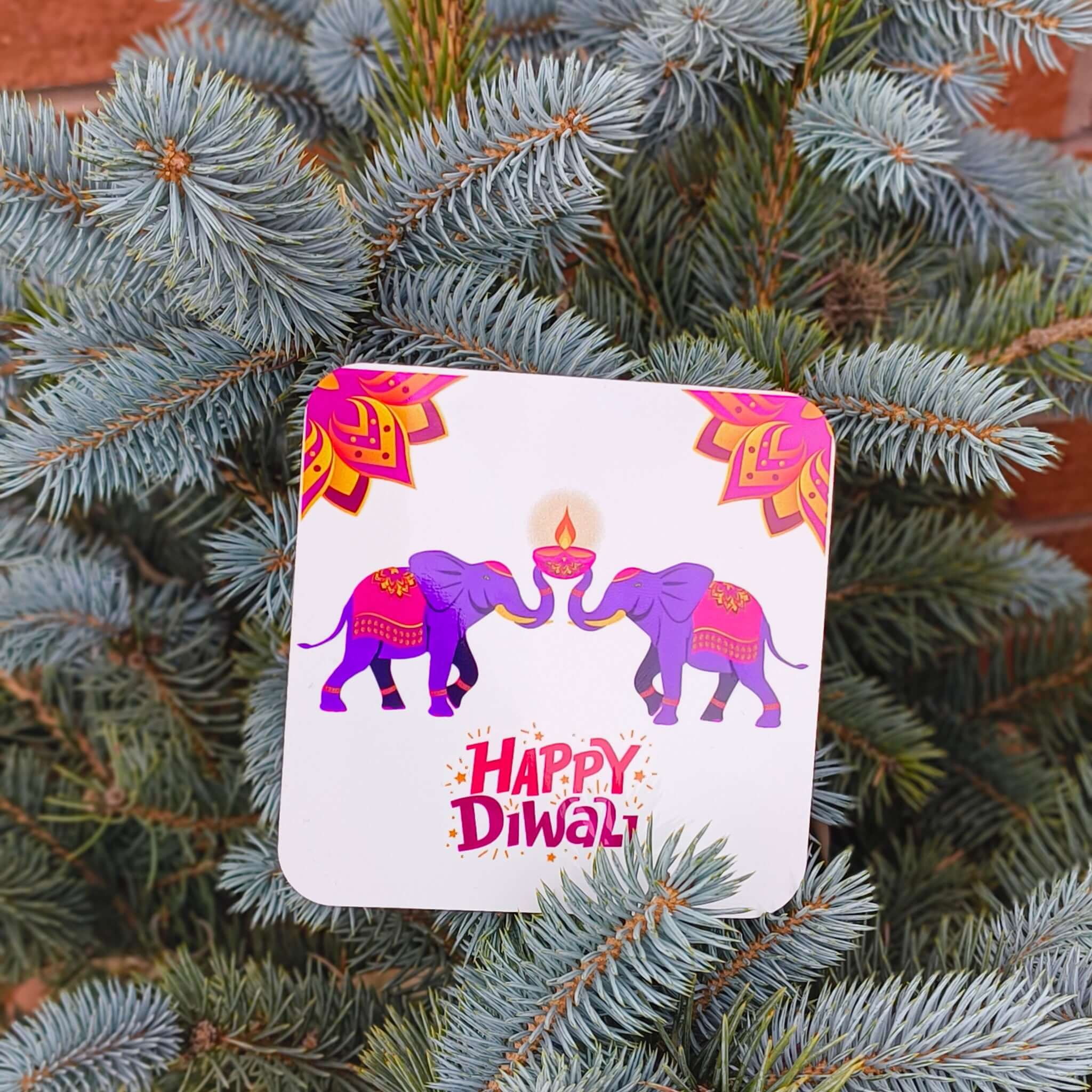 Happy Diwali coaster with two purple elephants and a central diya, surrounded by mandala motifs, photographed among blue spruce branches.