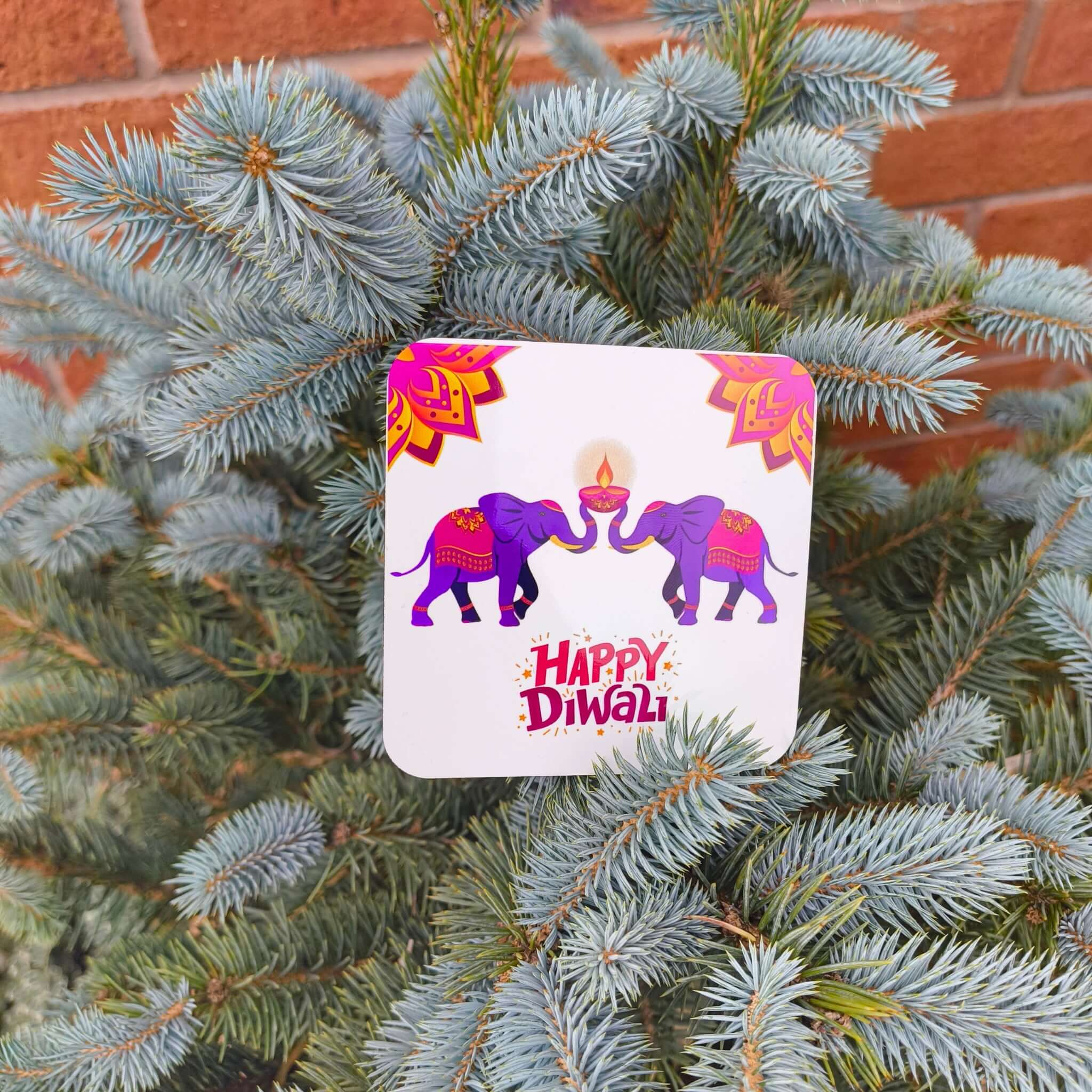 Happy Diwali coaster featuring two purple elephants holding a diya, displayed on a blue spruce tree outdoors against a brick wall.