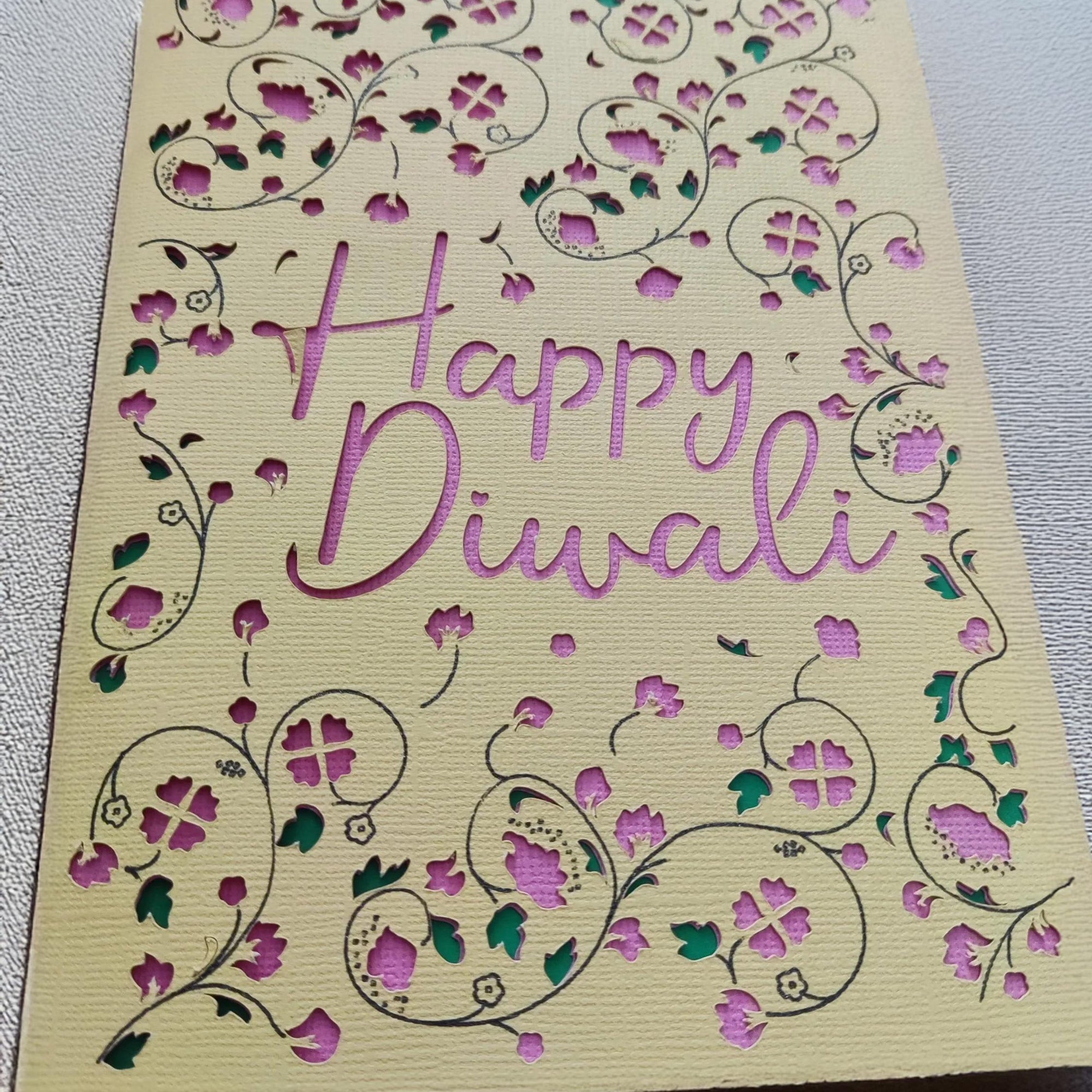 Close-up of a handmade Happy Diwali card featuring intricate floral cut-outs in pink and green against textured cream cardstock.