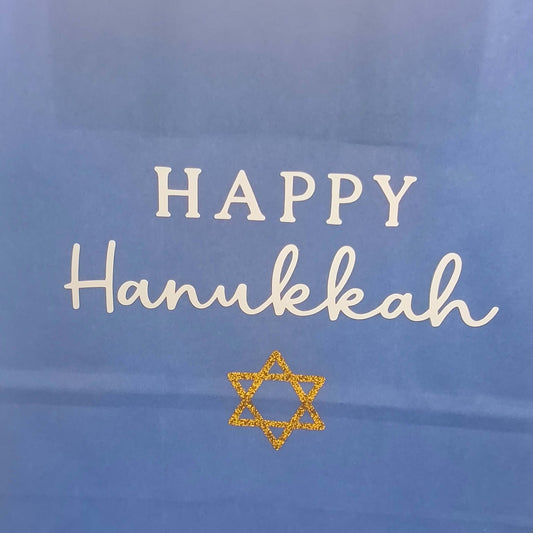 Close-up of a blue Hanukkah gift bag with white 'Happy Hanukkah' text and a gold glitter Star of David.