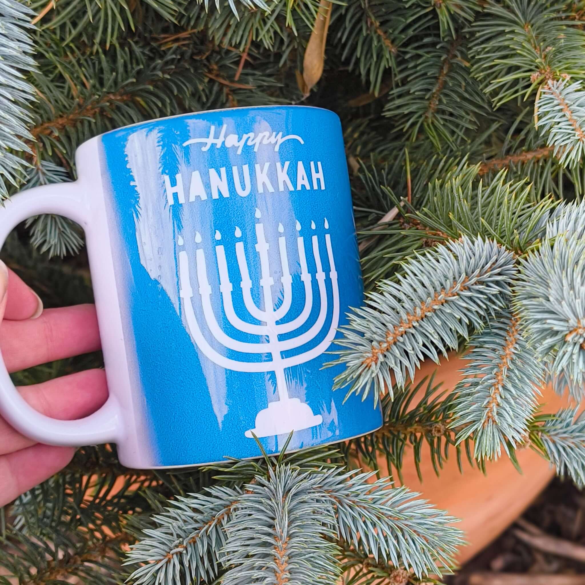 Happy Hanukkah mug displayed among green pine branches, featuring a blue design with a menorah.