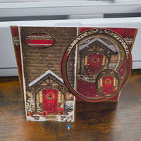 Handmade Christmas card featuring a cosy snow-covered cottage with a red front door and festive wreath, displayed on a wooden table.