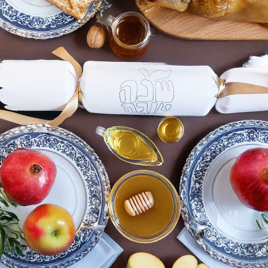 Festive table scene with a white Rosh Hashanah cracker at the centre, surrounded by honey, pomegranate, and matzah.