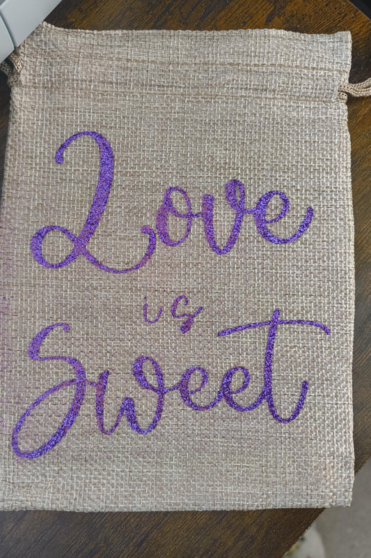 Rustic jute-style drawstring pouch featuring “Love is Sweet” in sparkling purple glitter. Compact size, ideal for sweets, favours, or small keepsakes.