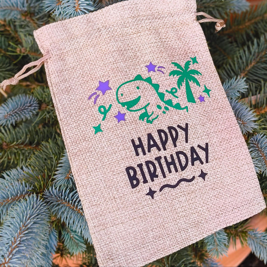 Large hessian Happy Birthday bag with dinosaur, palm tree, and stars design, photographed on blue spruce tree.