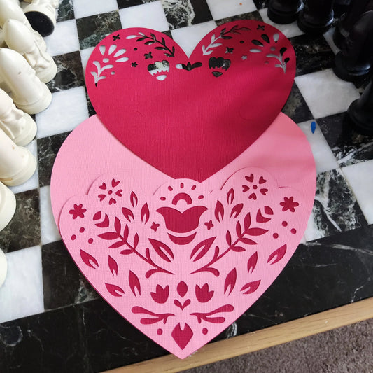 Layered heart papercut in progress, pink and red textured cardstock with floral cutouts, top-down angle on black and white chessboard surface, handmade romantic craft