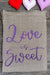 Small rustic hessian drawstring bag with “Love is Sweet” in purple glitter script. Shown with fabric hearts for scale. Natural woven texture adds a handmade charm.