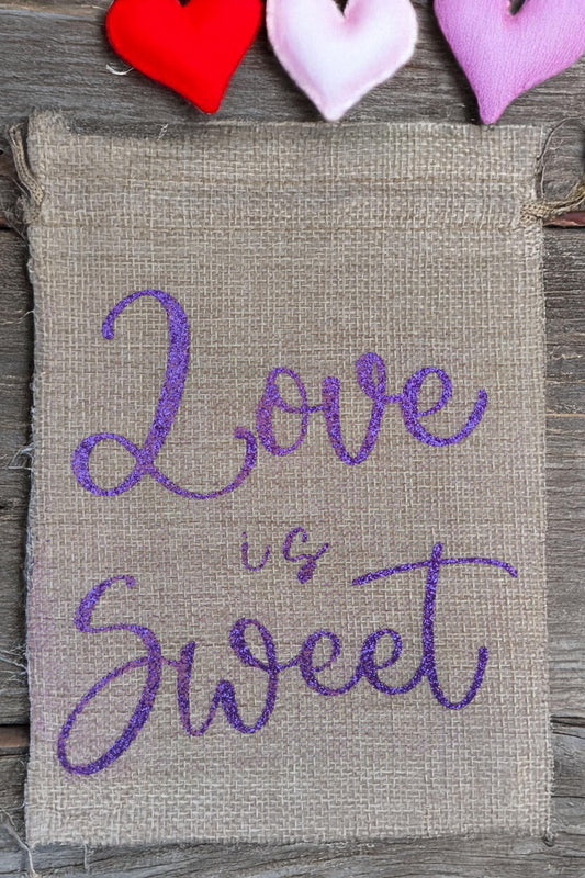 Small rustic hessian drawstring bag with “Love is Sweet” in purple glitter script. Shown with fabric hearts for scale. Natural woven texture adds a handmade charm.