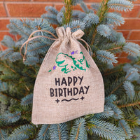 Medium hessian Happy Birthday gift bag with green dinosaur and purple stars design, displayed hanging on blue spruce tree branches.