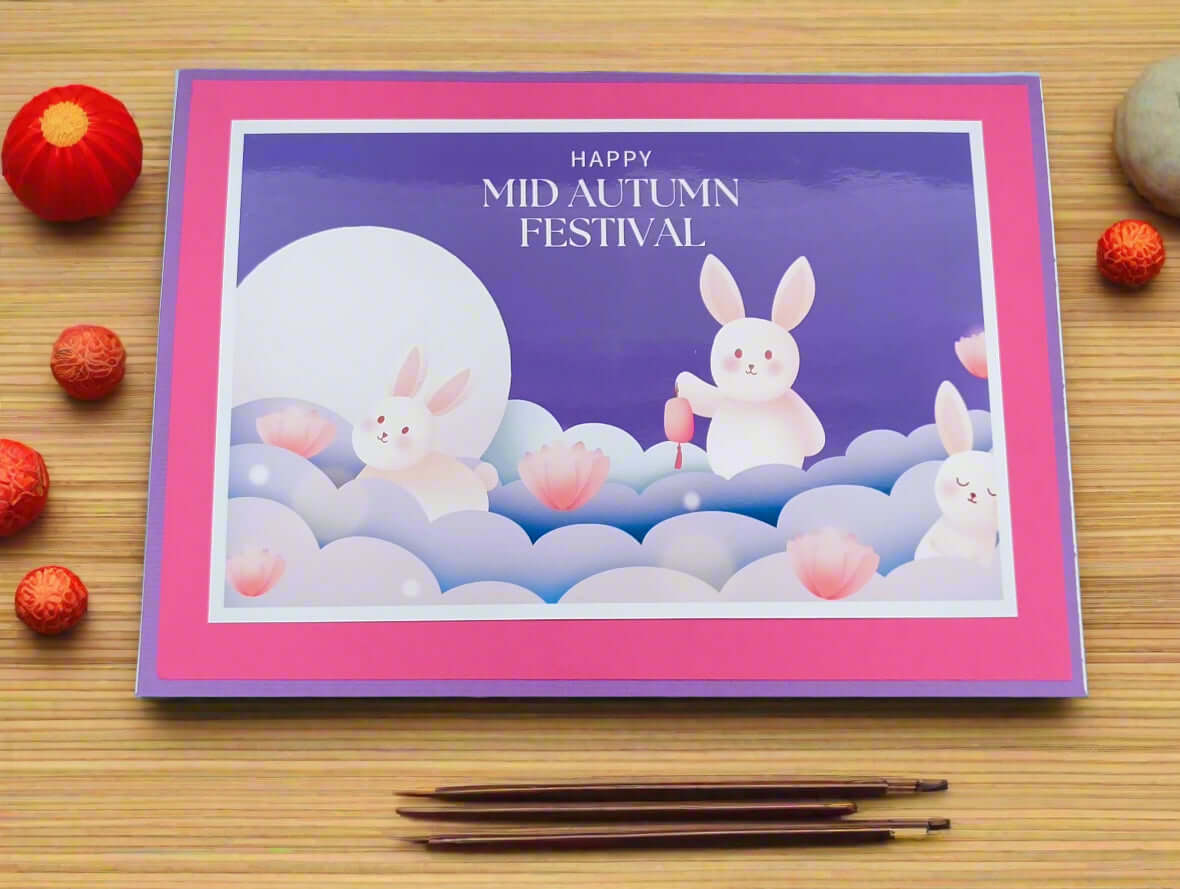Flat lay top-down view of a handmade Mid Autumn Festival card with cute moon rabbits and lotus flowers, styled on a bamboo mat with red lanterns, chopsticks, and decorative elements.