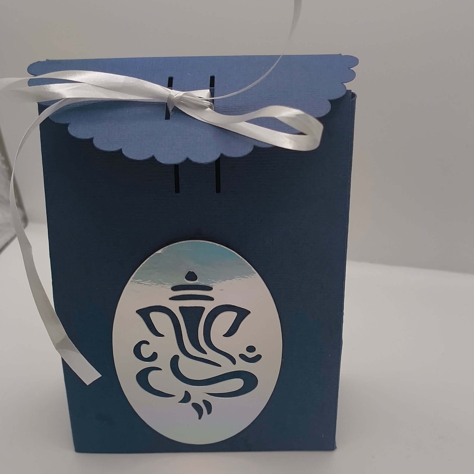 Handmade navy-blue Hindu gift box with scalloped lid, tied with white satin ribbon and silver Ganesh motif on the front, ideal for Diwali or wedding gifting.