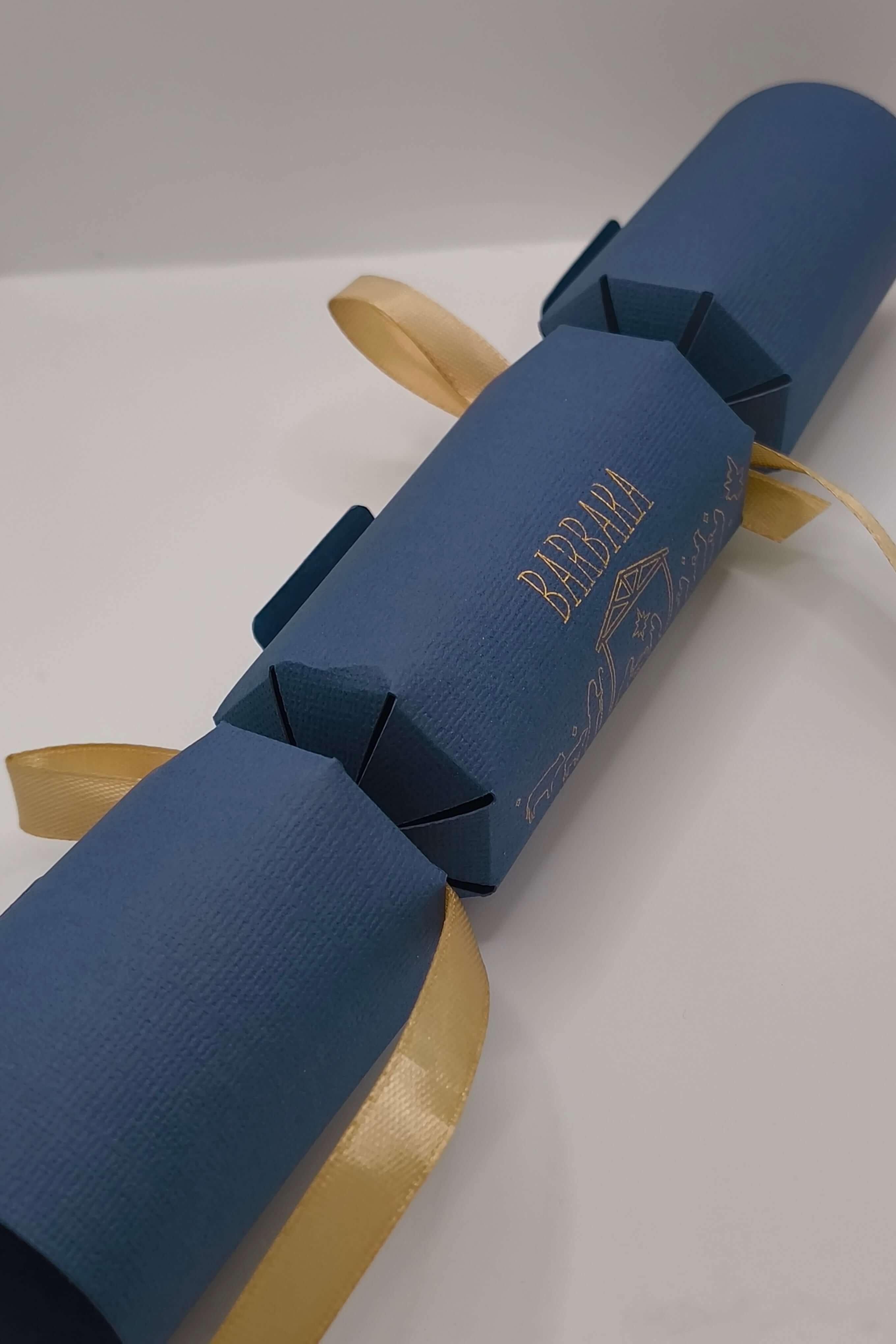 Luxury navy and gold handmade Christmas cracker shown at an angle, featuring Nativity design and satin ribbon ties.
