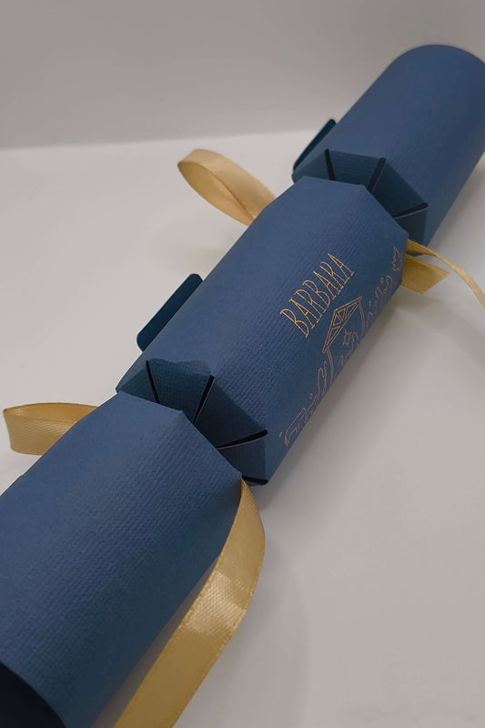 Luxury navy and gold handmade Christmas cracker shown at an angle, featuring Nativity design and satin ribbon ties.