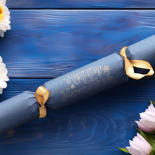 Handmade navy cracker with “Happy Hanukkah” hand-lettered in gold, finished with gold ribbon, styled with white and pink flowers on a blue wood background.