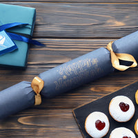 Hanukkah cracker in navy card with “Happy Hanukkah” lettering, gold ribbon ties, displayed on a rustic wood table with blue gift wrap and festive pastrie