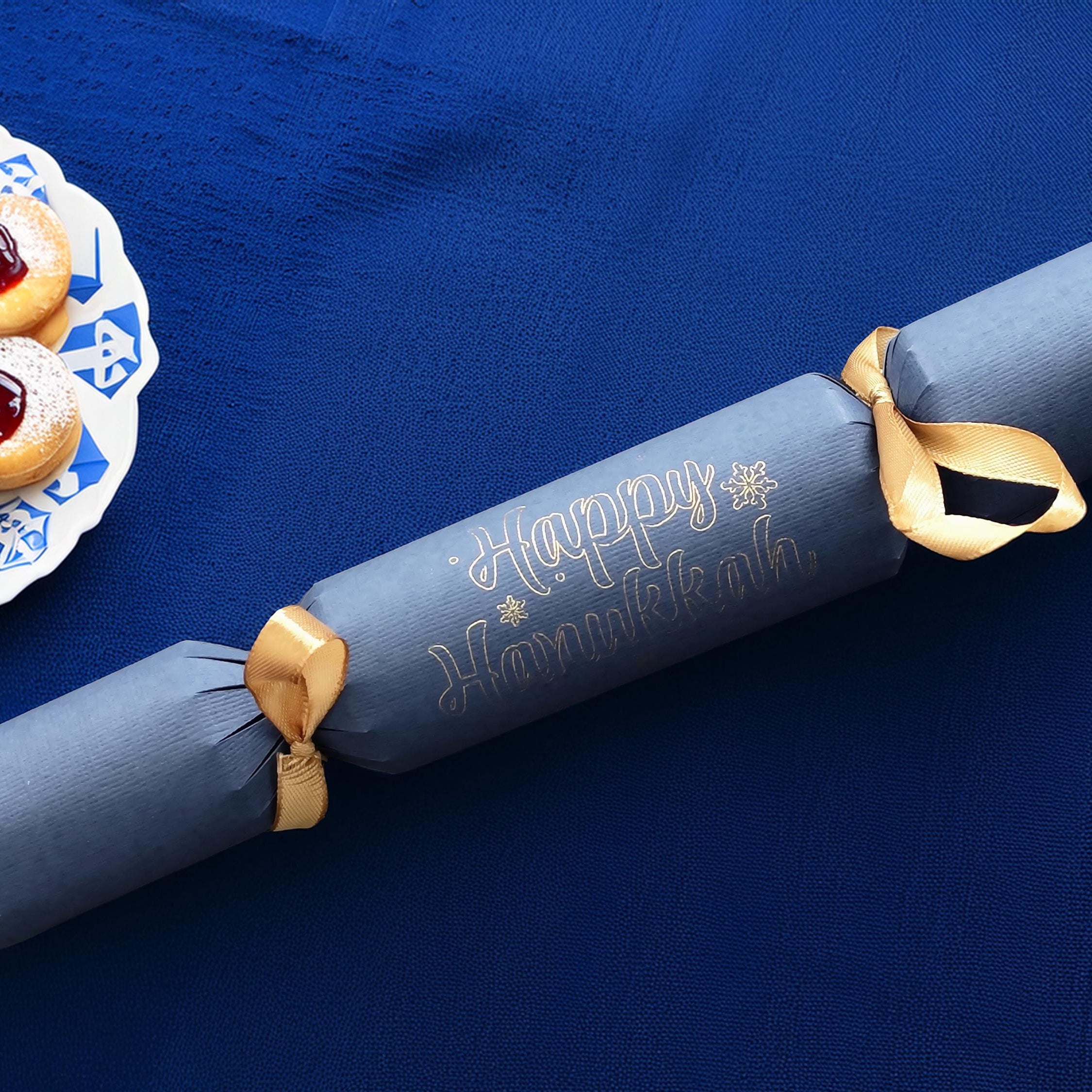 Navy blue cracker with “Happy Hanukkah” in silver pen, tied with gold ribbon, styled on a royal blue tablecloth beside jam-filled pastries.