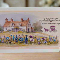 Front-facing image of a handmade New Home card featuring a charming countryside cottage with wildflowers and a purple horse-drawn carriage illustration, placed on a wooden surface with a soft, warm background.