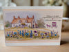 Front-facing image of a handmade New Home card featuring a charming countryside cottage with wildflowers and a purple horse-drawn carriage illustration, placed on a wooden surface with a soft, warm background.