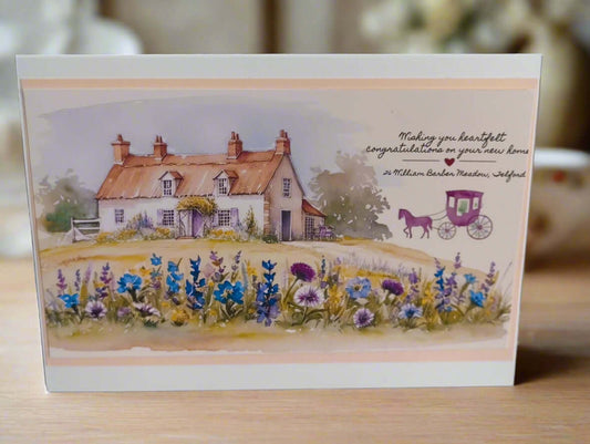 Front-facing image of a handmade New Home card featuring a charming countryside cottage with wildflowers and a purple horse-drawn carriage illustration, placed on a wooden surface with a soft, warm background.