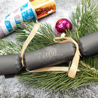 Handmade black New Year cracker tied with gold ribbon, featuring elegant âHappy New Yearâ lettering. Styled with greenery, baubles, and snow for a festive display.