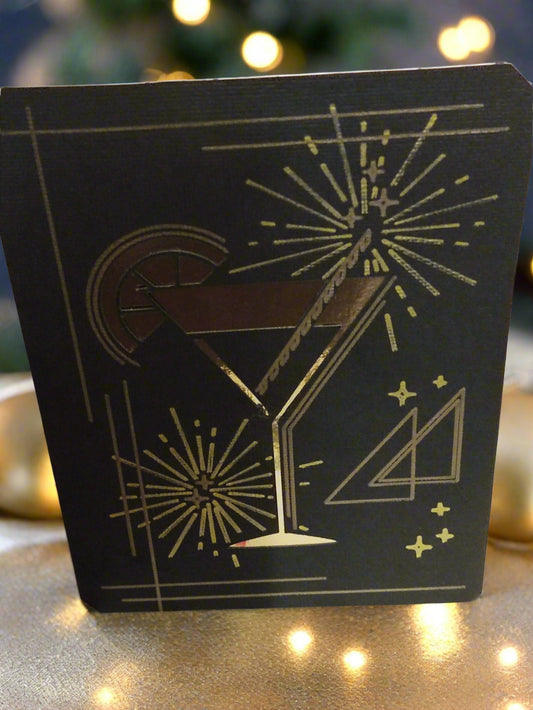 Foil cocktail glass New Year card, front angle, placed on a glittery gold surface with fairy lights and festive bokeh background