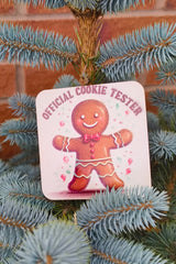 Close-up of festive âOfficial Cookie Testerâ gingerbread coaster with red bow tie design on a spruce tree.