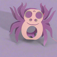 Pastel purple and pink spider-shaped lollipop holder made from textured cardstock, placed on a soft lilac background, perfect for Halloween party favours or handmade spooky gifts.