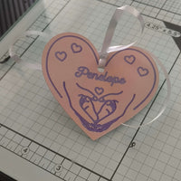 Angled view of handmade pink heart-shaped gift tag with purple heart and hand illustration, personalised with ‘Penelope’, tied with white satin ribbon, displayed on a craft cutting mat with gridlines.