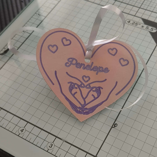 Angled view of handmade pink heart-shaped gift tag with purple heart and hand illustration, personalised with ‘Penelope’, tied with white satin ribbon, displayed on a craft cutting mat with gridlines.