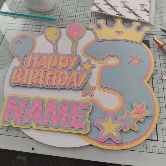 Personalised pastel cake topper for a 3rd birthday, shown from a top-down angle on a craft mat, featuring balloons, stars, crown, and “Happy Birthday” with space for a name.
