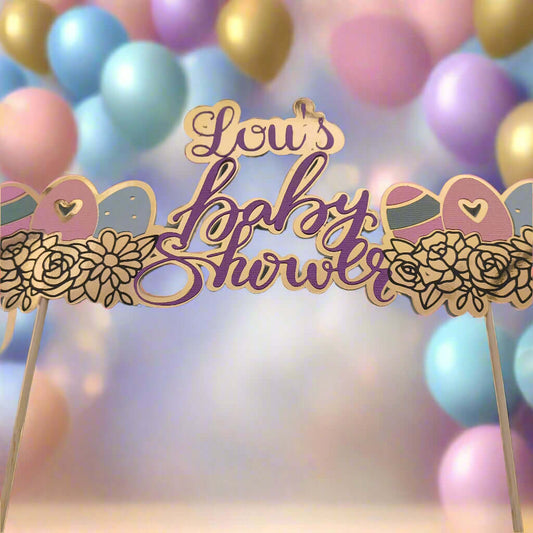 Gold and purple baby shower cake topper reading 'Lou's Baby Shower', decorated with floral and baby-themed elements. The topper is set against a soft-focus background of pastel balloons in blue, pink, lavender, and gold, creating a festive and dreamy atmosphere