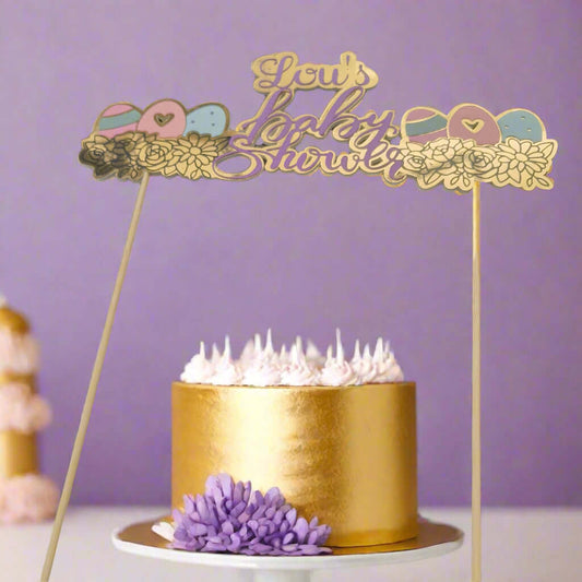 Gold and purple baby shower cake topper reading 'Lou's Baby Shower' placed on a metallic gold cake with white frosting and a purple flower. The soft lavender-purple background creates a festive and elegant baby shower setup.