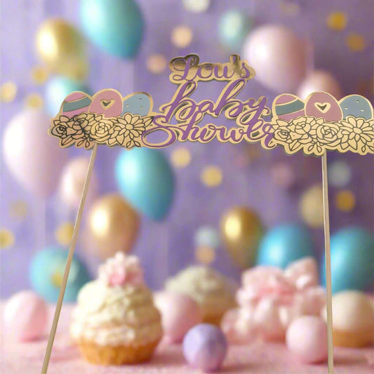 A pastel-themed baby shower table display with a decorative topper reading 'Lou's Baby Shower' in gold and purple script. The topper is adorned with floral and baby-themed designs and is held up by wooden sticks. In the background, there are soft-focus balloons in gold, pink, and blue tones, as well as cupcakes and pastel decorations.