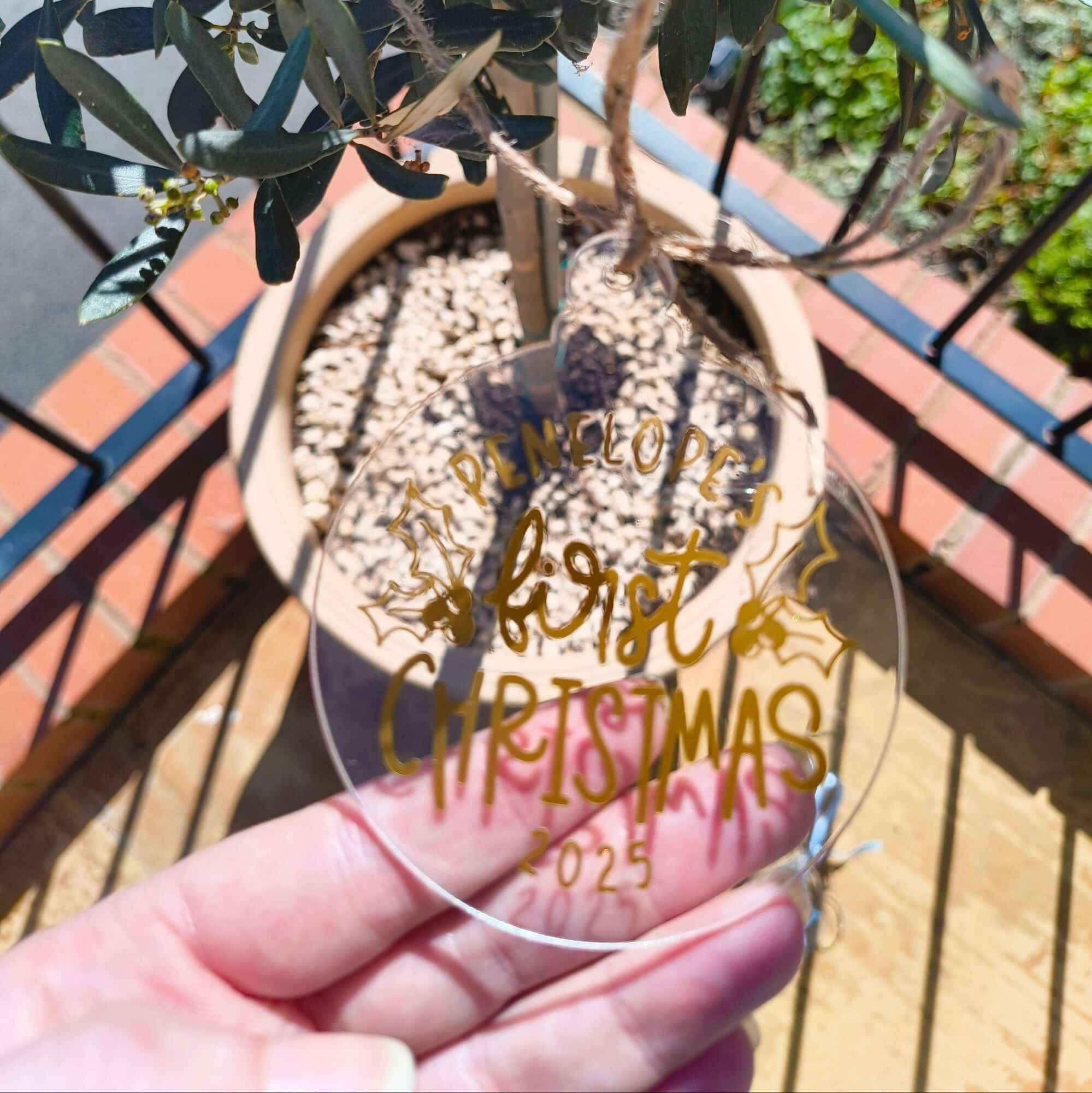Custom gold and clear acrylic first Christmas bauble held in hand with potted plant in background.