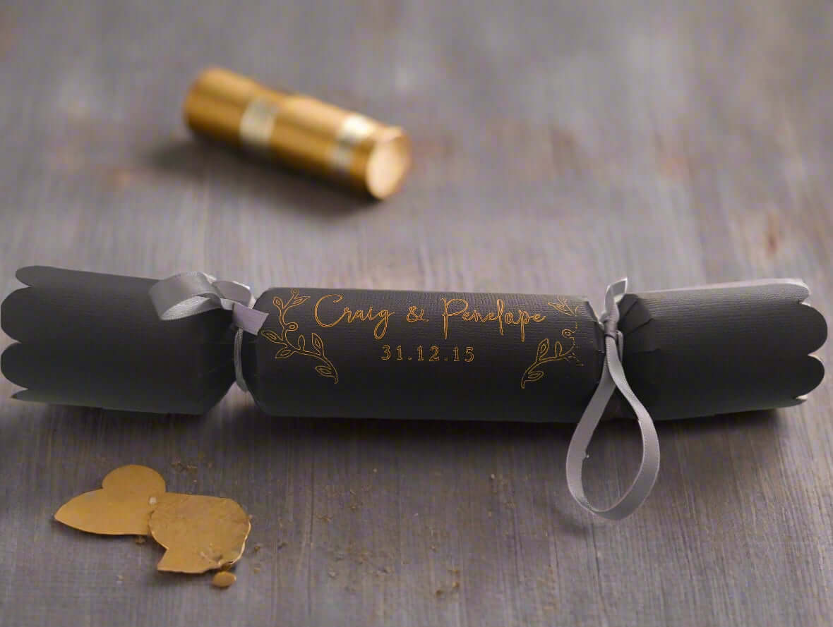 Close-up of black cracker with gold foil text “Craig & Penelope” and date, decorated with silver ribbon and elegant leaf motifs.