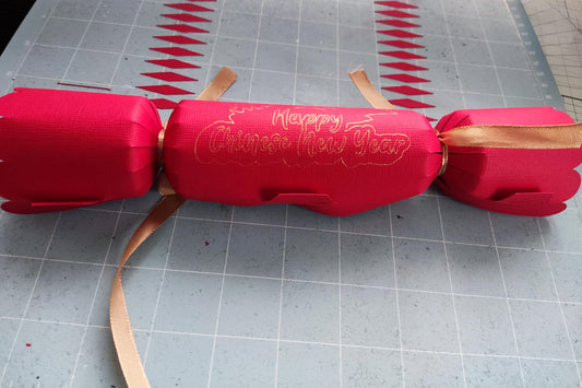 Red cracker with gold foil “Happy Chinese New Year” message, tied with gold ribbon, placed on a craft mat background alongside paper cut-outs.