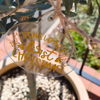 Custom acrylic Christmas ornament with gold lettering reading "Penelope’s First Christmas 2025" hanging outdoors.