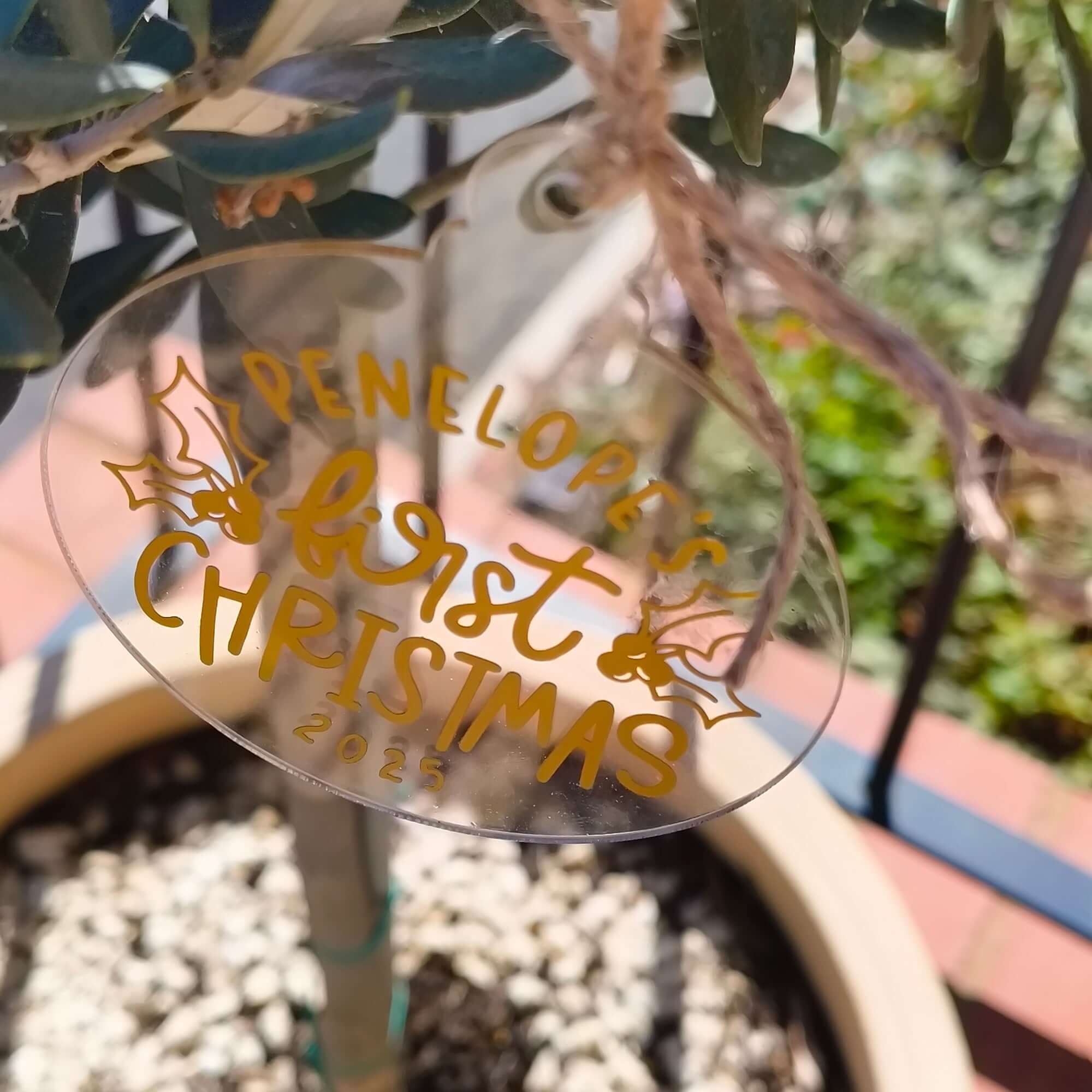 Custom acrylic Christmas ornament with gold lettering reading "Penelope’s First Christmas 2025" hanging outdoors.