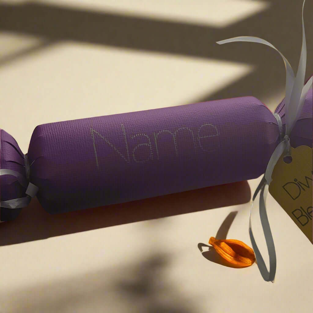 Personalised purple Diwali cracker with white ribbon and 'Diwali Blessings' tag, featuring a space for a name and a small orange diya decoration on a sunlit background.