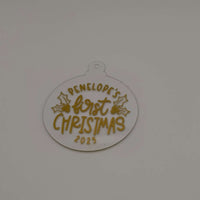 Clear acrylic bauble with gold lettering reading "Penelope’s First Christmas 2025" on plain background.